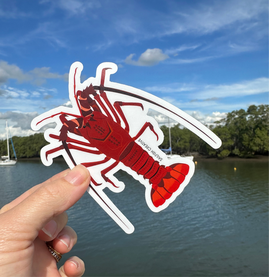 Hand holding a Crayfish Rock Lobster sticker with a river and trees in the background under a blue sky