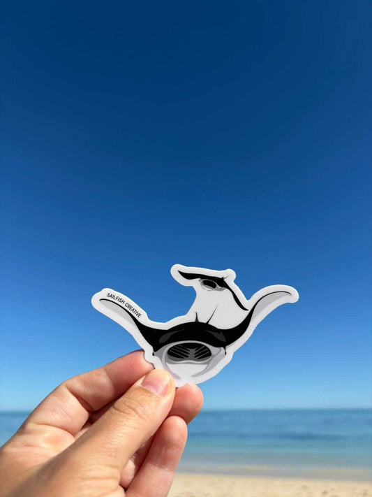 Hand holding a black and white Mantaray sticker against a blue sky and beach background