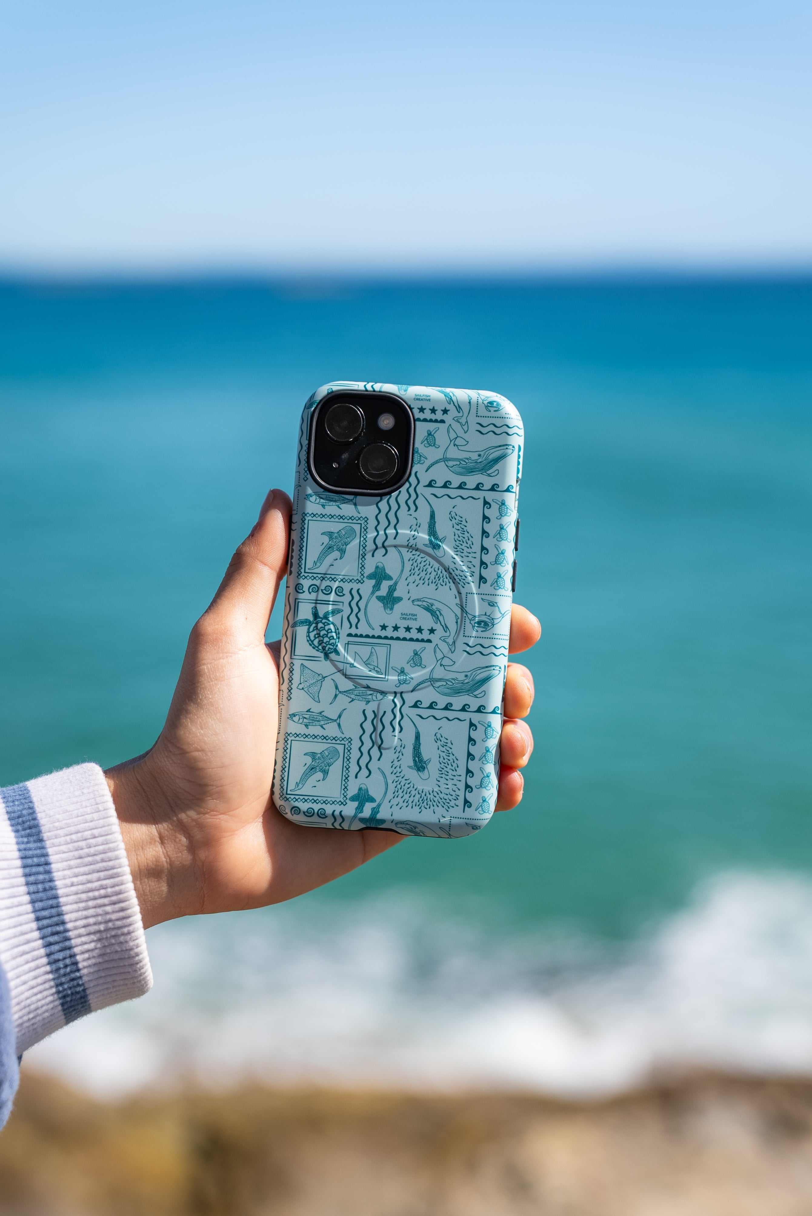 iPhone Phone Case Busy Print Teal with ocean animal design held by hand near beach