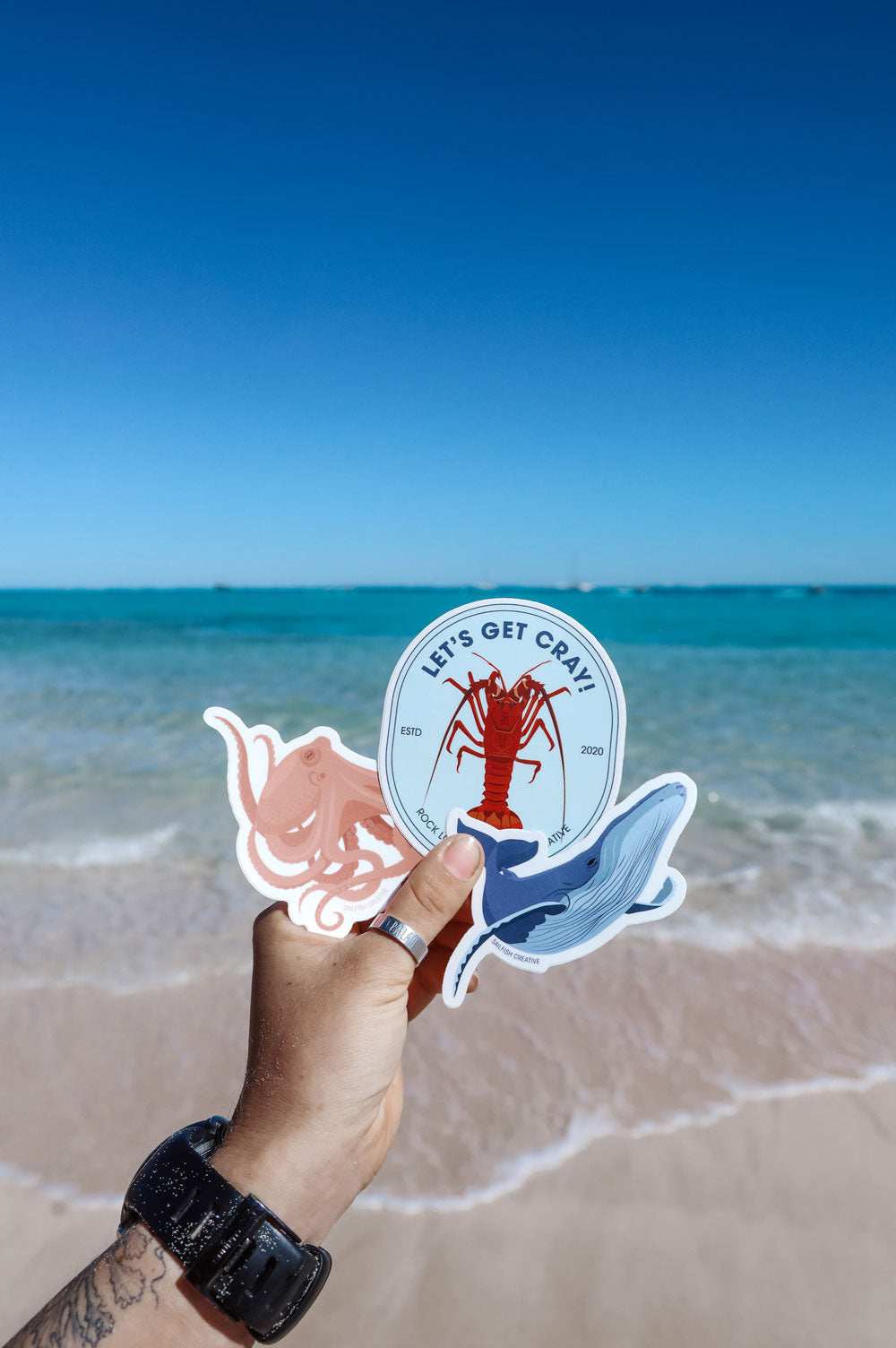 Hand holding Crayfish Pun sticker by the beach with ocean background