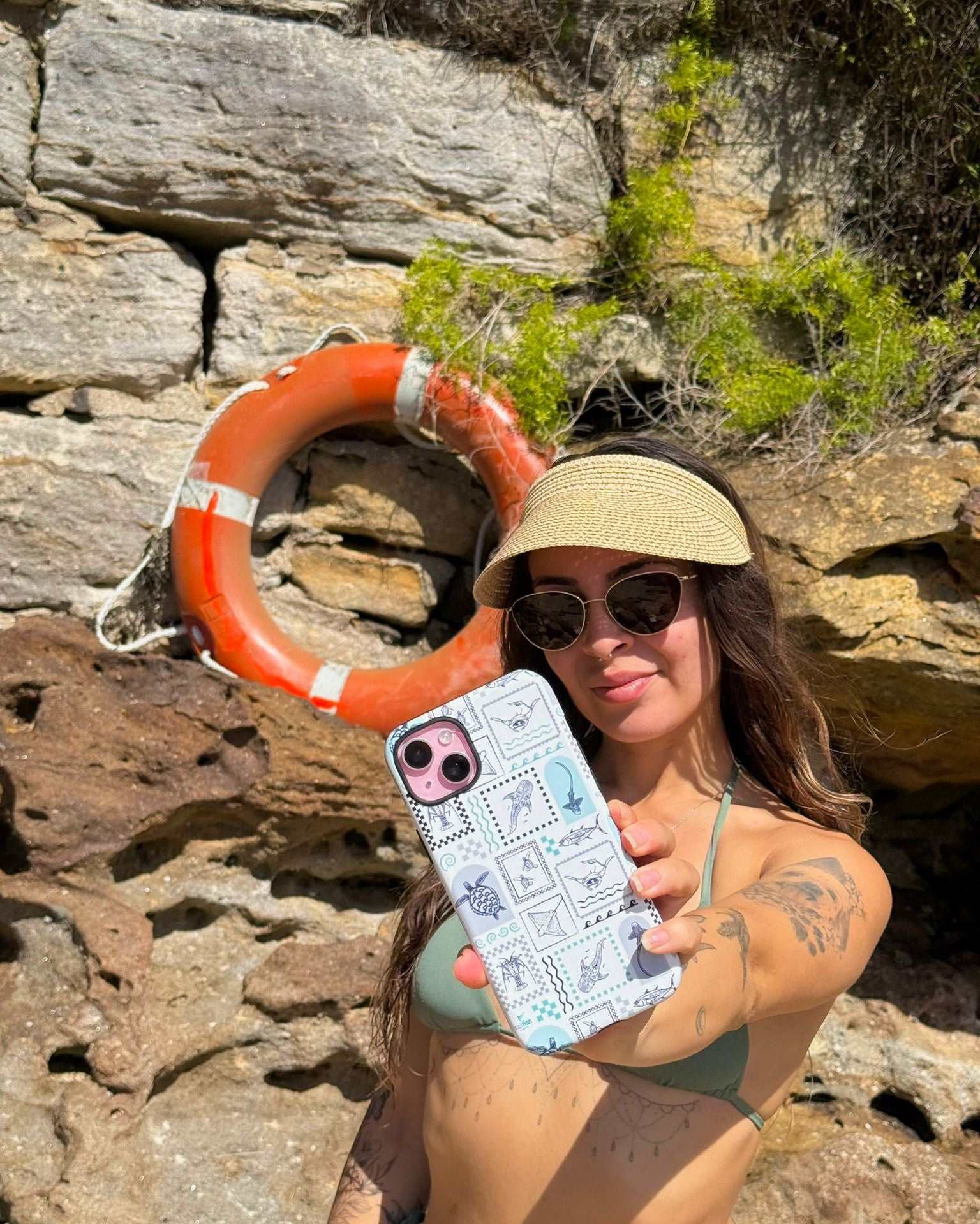 Sammy holding a arches print case on iphone, wearing sunglasses and a visor, standing on a rocky beach in Sydney.