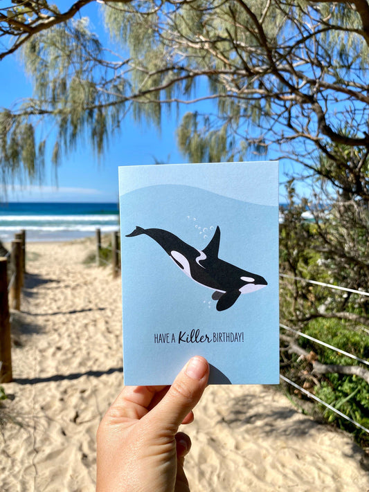 Birthday card featuring a killer whale orca design held at a beach pathway with trees and ocean background