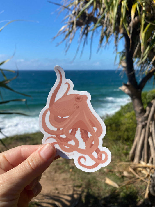 Hand holding an octopus sticker by the ocean with trees and blue sky in the background