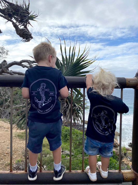 Two kids wearing Feelin' Nauti Kids T-Shirt with anchor design standing by a wooden fence near the ocean