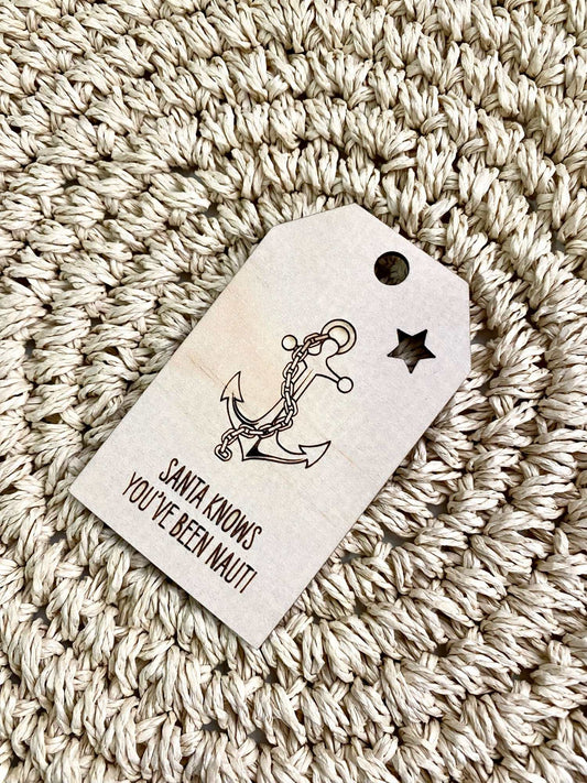 Wooden Christmas swing tag with anchor and chain design and "Santa knows you've been nauti" text on woven background