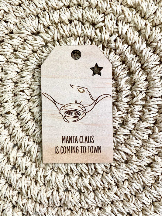 Wooden Christmas swing tag with manta ray design and "Manta Claus is coming to town" text on plywood background