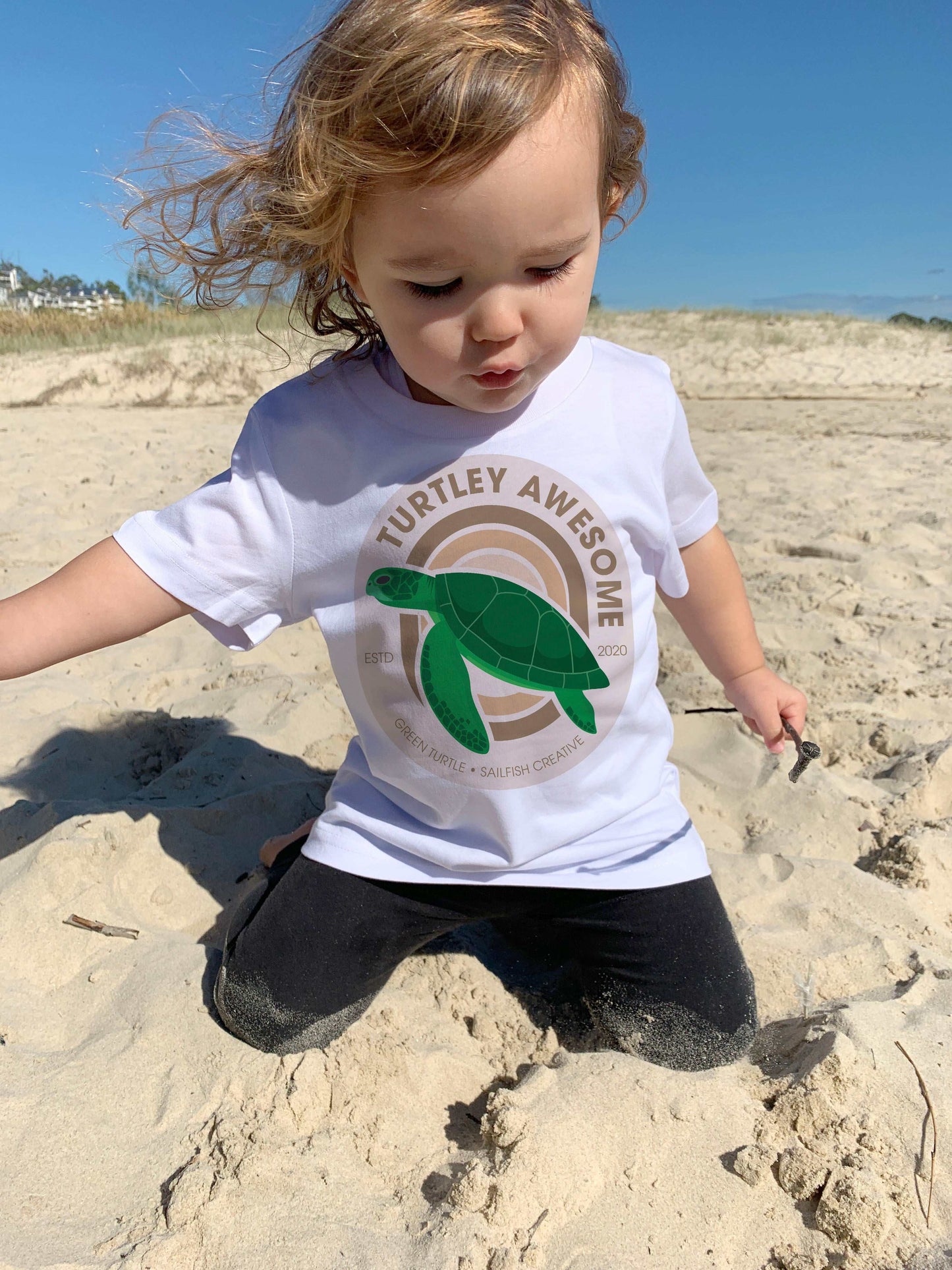 Toddler wearing white "Turtley Awesome" kids t-shirt with green turtle graphic playing on sandy beach