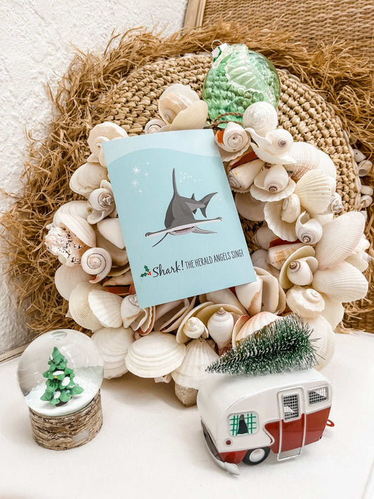 Christmas card featuring humorous hammerhead shark illustration surrounded by seashell wreath and festive decorations