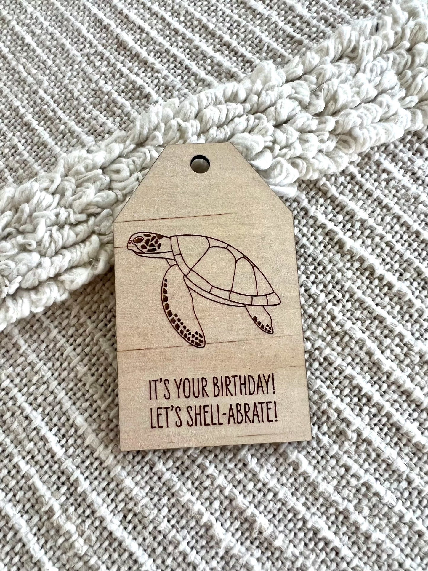 Wooden birthday gift tag with green turtle design and "It's your birthday! Let's shell-abrate!" text on textured fabric background