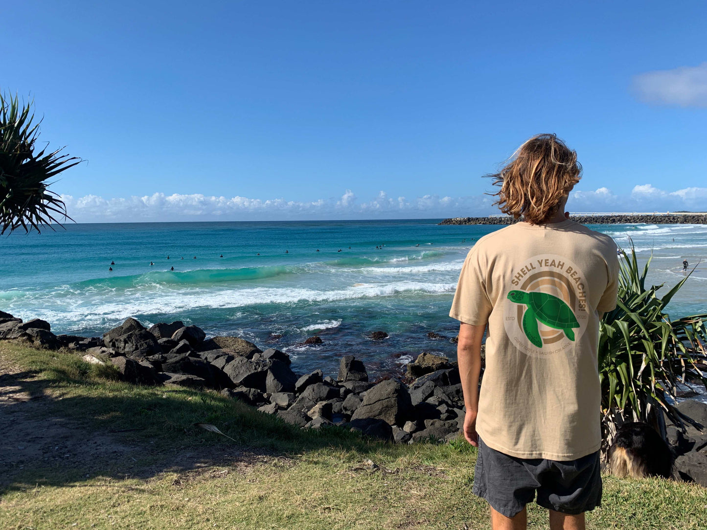 Man wearing Shell Yeah Beaches t-shirt with green turtle graphic standing by the beach ocean view