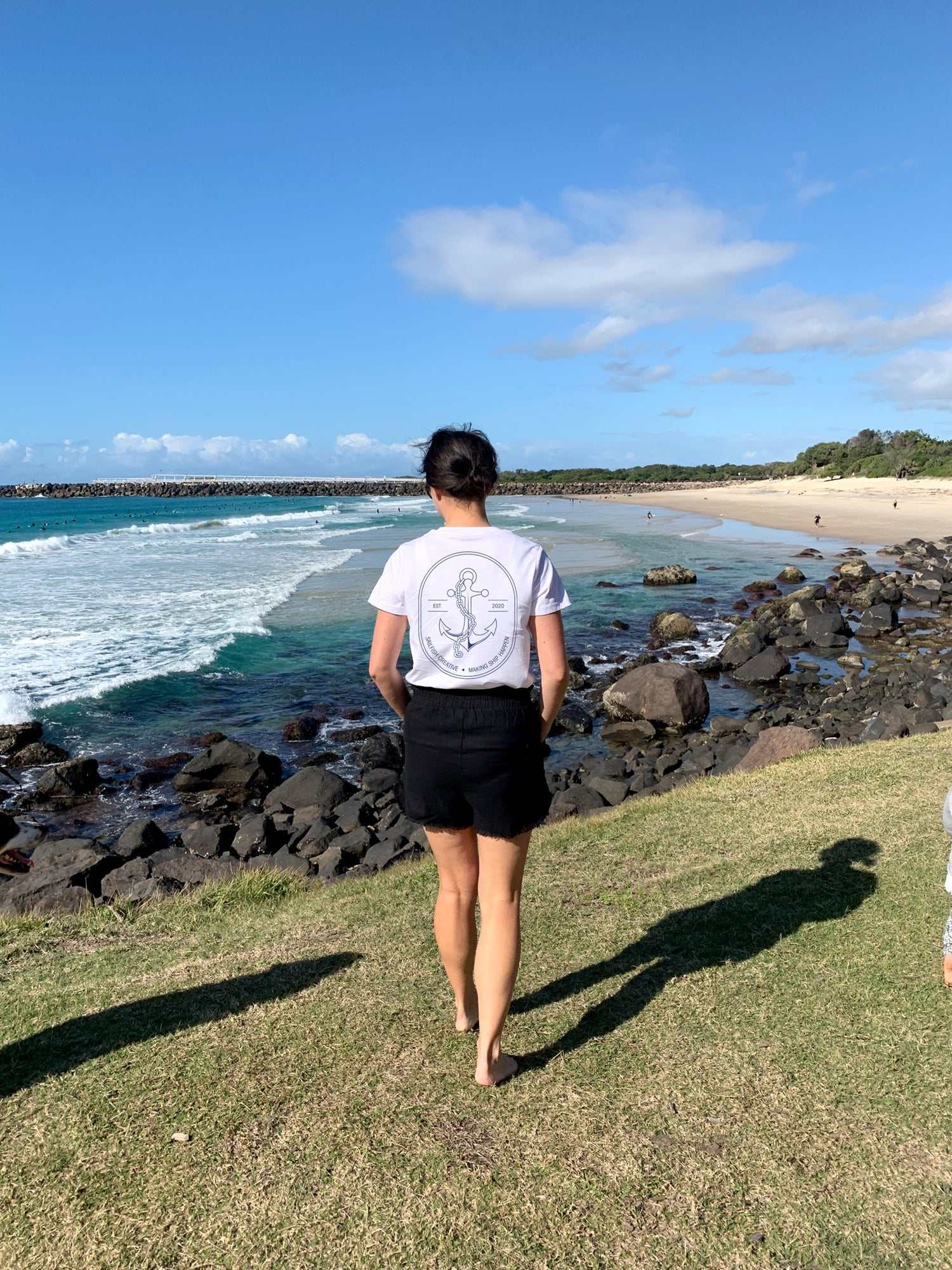 Woman wearing Making Ship Happen white t-shirt by the beach with ocean and rocks in the background