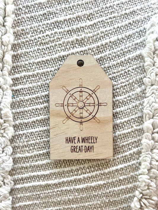 Wooden gift tag with ships wheel design and "Have a wheely great day" message on textured fabric background