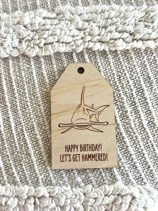 Wooden birthday gift tag with engraved hammerhead shark and humorous birthday message on textured fabric background