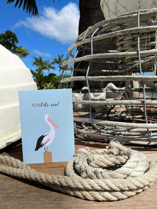 Pelican greeting card with cartoon pelican and "Yes You Peli-can!" text on blue background displayed outdoors near nautical rope and fishing gear