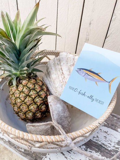 Greeting card with tuna fish illustration beside pineapple and seashells in a basket