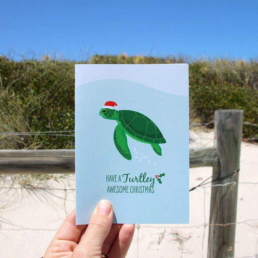 Hand holding Christmas card with green turtle wearing Santa hat at the beach